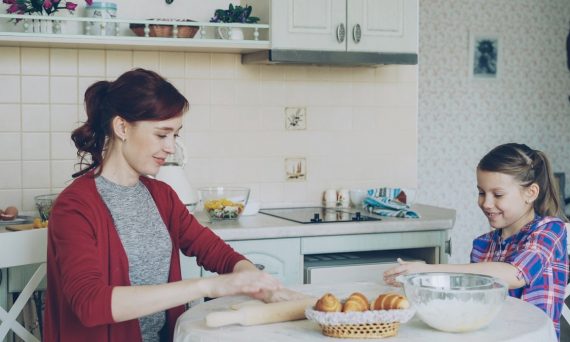 Young mother rolling dough and talking to her little cute daughter while cooking in the kitchen on weekend. Family, food, home and people concept