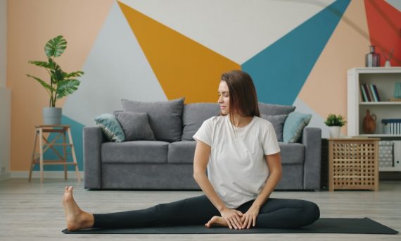 Attractive girl in leggings and T-shirt is doing stretching exercises on yoga mat relaxing on floor at home busy with practise. Youth, lifestyle and health concept.
