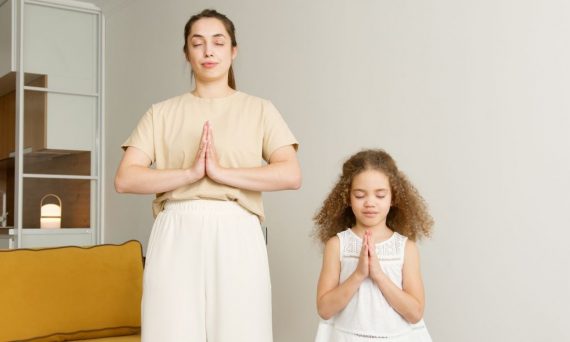 A mother and daughter practicing meditation and prayer indoors with eyes closed.